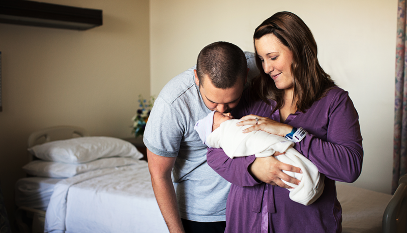 A smiling woman holds her newborn while her husband kisses it