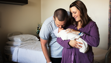 A smiling woman holds her newborn while her husband kisses it on the forehead.